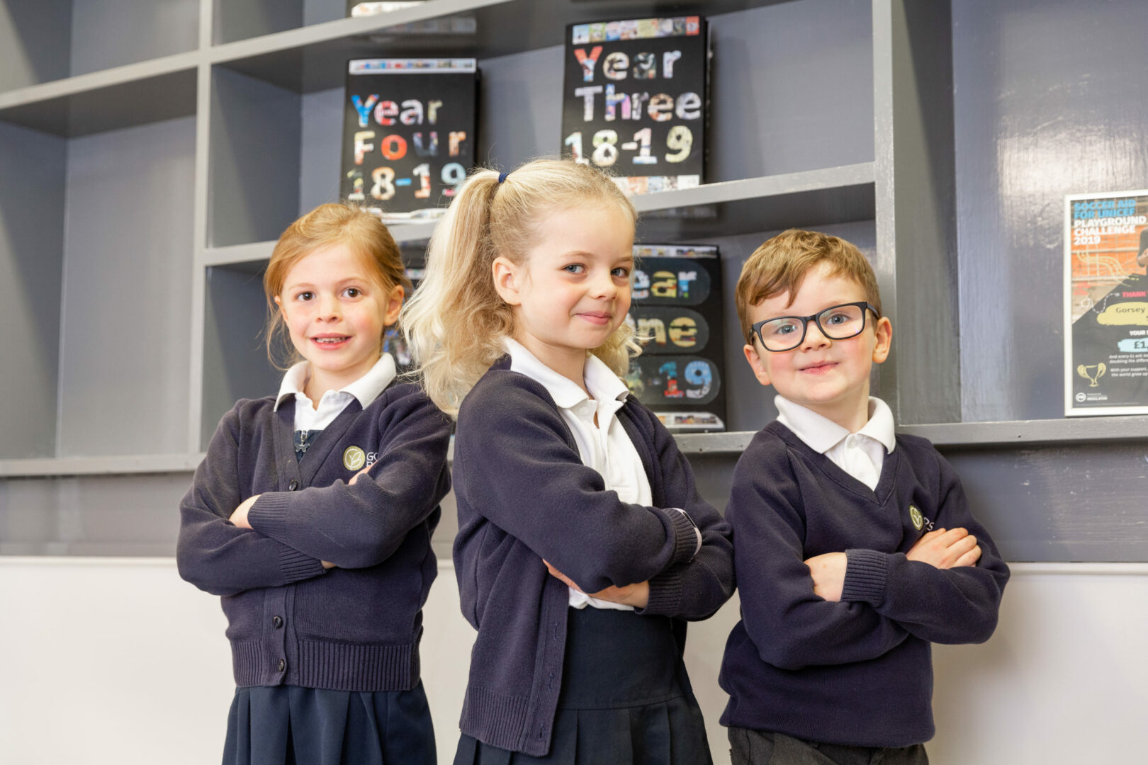 Three Gorsey Bank Primary School pupils stand smiling and crossing their arms.